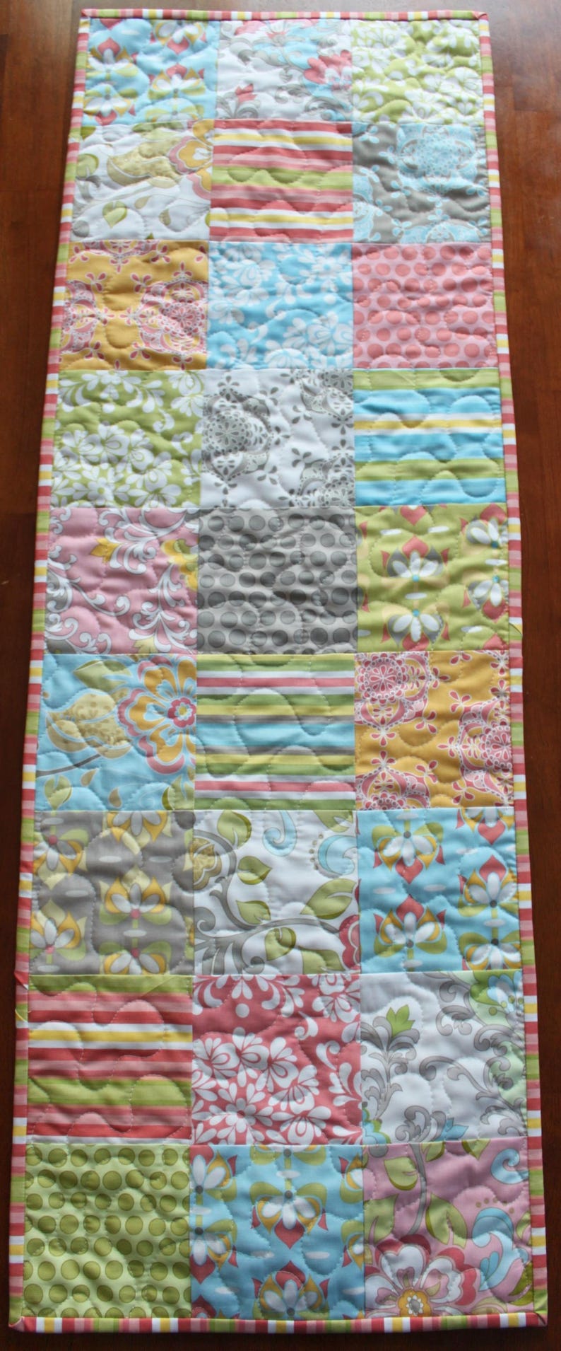 May include: A patchwork table runner with a variety of floral and geometric patterns in shades of pink, green, blue, and gray. The runner has a striped border.