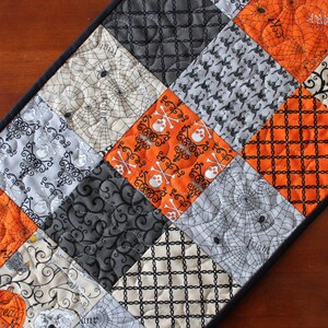 May include: A Halloween table runner with a patchwork design featuring various spooky patterns in black, white, and orange. The patterns include skulls and crossbones, bats, spiders, and webs.