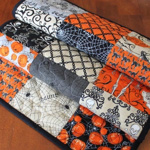 May include: A stack of Halloween-themed quilted placemats. Each placemat features a patchwork design with various patterns, including pumpkins, black cats, spiderwebs, and skulls. The color palette includes orange, black, white, and gray, with a dark blue border.