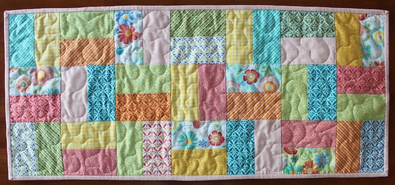 May include: A rectangular patchwork table runner with a variety of colourful fabric squares. The squares feature floral, geometric, and textured patterns in shades of pink, blue, green, yellow, and orange. The runner is approximately 91 cm long and 30 cm wide.