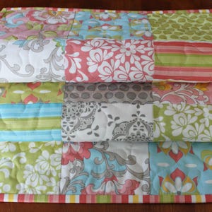 May include: A quilted table runner with a patchwork design featuring various floral patterns in shades of pink, green, gray, and blue. The runner has a scalloped edge and a pink, green, and yellow trim.