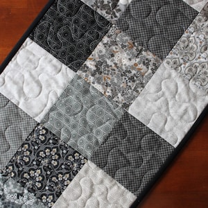 May include: A quilted table runner featuring a patchwork design in shades of gray, black, and white. The squares showcase various patterns, including floral, geometric, and solid textures. The runner has a black border and is placed on a wooden surface.