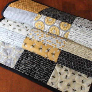 May include: A folded, quilted table runner with a bee theme. The runner features a patchwork design with various fabrics in yellow, gray, black, and white. Some squares have bee and honeycomb patterns, while others have script or floral designs.