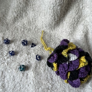 May include: A crocheted dice bag in a scale pattern, featuring purple, black, yellow, and white colors. Several polyhedral dice in shades of blue and green are scattered nearby. The bag has a yellow drawstring.