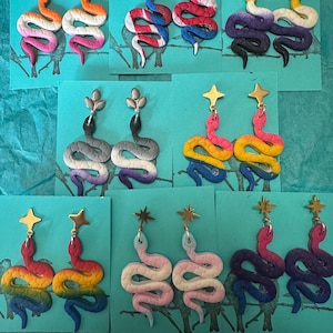 May include: Eight pairs of dangling snake earrings in various gradient color combinations. Each snake features a textured surface and is attached to a gold or silver-toned star or leaf-shaped stud. The earrings are displayed on individual teal cards.