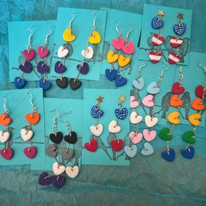 May include: A collection of multiple pairs of dangle earrings featuring colorful heart-shaped charms suspended from silver-toned metal chains. Each pair is attached to a teal card with a bird silhouette print, showcasing various color combinations.