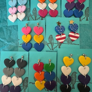 May include: Eight pairs of dangling heart-shaped earrings displayed on teal cards. Each pair features three stacked hearts in various color combinations, including pastels, primary colors, and patterns like polka dots and stripes, with metal posts.