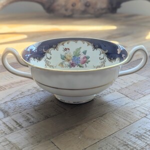 May include: A vintage soup bowl with a white base, navy blue rim, and gold trim. The bowl has two handles and a floral design in the centre. The floral design includes pink, blue, and yellow flowers. The bowl is a classic design.