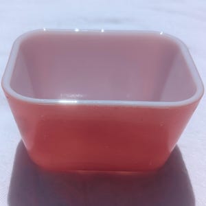 May include: A square-shaped bowl made of opaque pink glass with rounded corners and a smooth, glossy finish. The bowl features a wide, open top and slightly tapered sides that extend down to a flat base, presenting a simple, functional design.