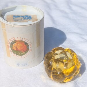 May include: A round, amber-colored glass candle holder with a textured, bubble-like surface, accompanied by a cylindrical white cardboard box. The box features a label with an illustration of a cloudberry and the text Nordkalottens Rubusljus.