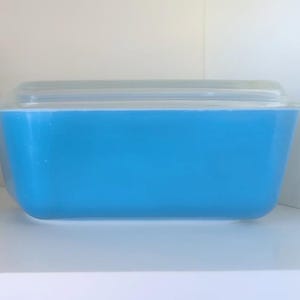 Op de afbeelding: Twee vintage Pyrex-ovenschalen met doorzichtige deksels. Eén is een oranje kom met een wit bloemmotief. De andere is een rechthoekige, lichtblauwe schaal. Beide staan op een witte plank.