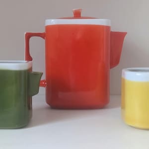 Op de afbeelding: Een set van drie keramische keukenartikelen: een rode koffiepot met een deksel, een groene mok en een gele mok. Elk stuk heeft een witte rand. De koffiepot heeft een handvat en een tuit.