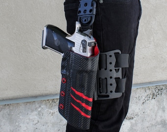 Puede incluir: Una funda de pierna táctica negra y roja con una pistola plateada. La funda tiene un patrón de fibra de carbono y detalles rojos.