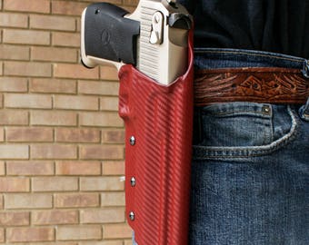 Puede incluir: Una funda de cuero roja con una pistola plateada en su interior. La funda está sujeta a un cinturón de cuero marrón.
