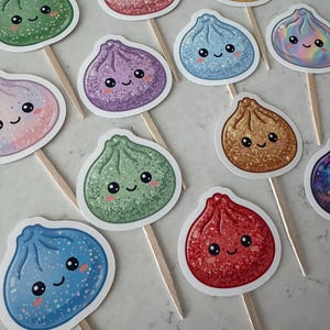 May include: A collection of cupcake toppers featuring various dumpling-shaped illustrations with smiling faces. Each topper is attached to a wooden pick and displays a unique color, including green, pink, yellow, blue, purple, and red, with glittery textures.