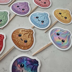 May include: A collection of cupcake toppers featuring various dumpling-shaped characters with smiling faces. Each topper is attached to a wooden pick and displays a unique finish, including glittery, holographic, and galaxy-patterned designs in colors like blue, purple, red, green, and gold.