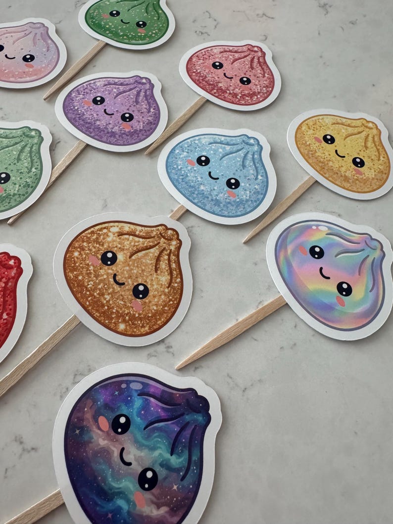 May include: A collection of cupcake toppers featuring various dumpling-shaped characters with smiling faces. Each topper is attached to a wooden pick and displays a unique finish, including glittery, holographic, and galaxy-patterned designs in colors like blue, purple, red, green, and gold.