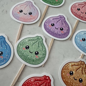 May include: A collection of decorative food toppers featuring various colorful, glittery dumpling illustrations with smiling faces. Each dumpling is attached to a wooden pick, showcasing shades of green, purple, pink, blue, and brown with sparkling details.
