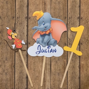 May include: Three cake toppers featuring a gray elephant with a yellow hat, a mouse wearing a red and blue uniform, and a yellow number one. The elephant and mouse toppers are on sticks, and the number one topper is on a stick. The elephant topper has a cloud with the name "Julian" written on it.