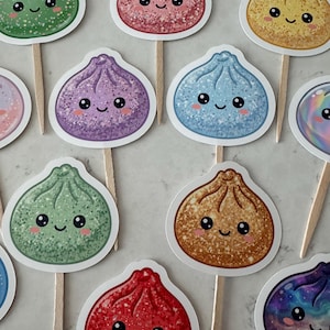 May include: A collection of cupcake toppers featuring various dumpling-shaped characters with glittery textures in green, pink, yellow, purple, blue, brown, and red. Each dumpling has a smiling face with black eyes and pink cheeks on a wooden stick.