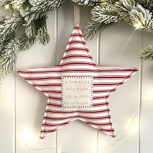 May include: A star-shaped fabric ornament featuring a red and white striped pattern. A square off-white patch is stitched to the centre with the text: Silent night, Holy night, All is calm, All is bright. The ornament hangs from a fabric loop.