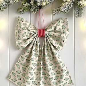 May include: A large fabric bow decoration featuring a repeating pattern of green pine trees on a cream-coloured background. The centre of the bow is cinched with a red and white textured fabric band, and it includes a thin red string for hanging.
