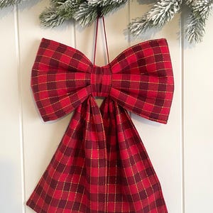 May include: A decorative bow ornament featuring a red and dark red plaid pattern with thin gold metallic thread accents. The bow has two large loops and two long, hanging tails, suspended by a thin red string for hanging purposes.