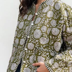 Peut inclure: Veste matelassée vert olive avec un motif floral blanc. La veste a un col mandarin, des boutons blancs et deux poches avant. Les bords sont ornés d'un motif rayé noir et blanc. Un vêtement d'extérieur élégant.