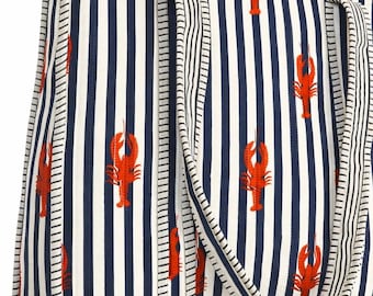 May include: A tote bag with a nautical theme, featuring a pattern of red lobsters on a white and navy blue striped background. The bag has matching striped handles and trim, and is sitting on a light wooden table.