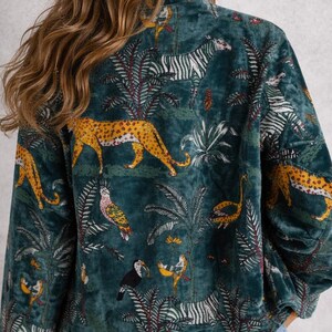 May include: Teal velvet jacket with a jungle-themed print. The design features zebras, leopards, birds, and tropical foliage. The jacket has a high collar and long sleeves, and is paired with blue jeans.