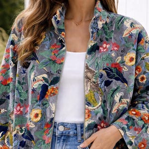 Peut inclure: Veste grise à imprimé floral avec un col montant et une fermeture éclair. La veste présente un motif coloré de fleurs et de feuilles dans les tons de rouge, jaune, bleu et vert. Le modèle porte un jean bleu.