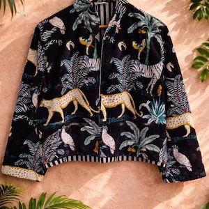 K&ouml;nnte beinhalten: Schwarze Jacke mit einem lebendigen Dschungel-Print, der Leoparden, Zebras und Papageien zwischen Palmen zeigt. Die Jacke, die an einem Holzb&uuml;gel h&auml;ngt, hat einen Kragen. Der Saum und die Manschetten haben ein Streifenmuster.