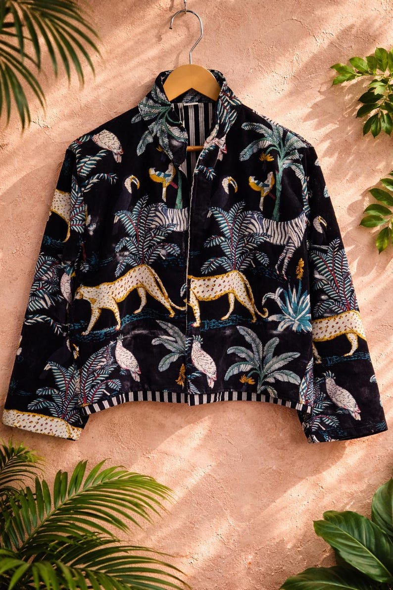 K&ouml;nnte beinhalten: Schwarze Jacke mit einem lebendigen Dschungel-Print, der Leoparden, Zebras und Papageien zwischen Palmen zeigt. Die Jacke, die an einem Holzb&uuml;gel h&auml;ngt, hat einen Kragen. Der Saum und die Manschetten haben ein Streifenmuster.
