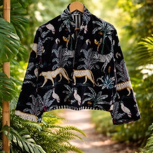 K&ouml;nnte beinhalten: Schwarze Samtjacke mit Dschungel-Print. Das Muster zeigt Leoparden, Zebras, V&ouml;gel und Palmen. Die Jacke h&auml;ngt an einem Holzb&uuml;gel vor dem Hintergrund &uuml;ppigen Gr&uuml;ns.
