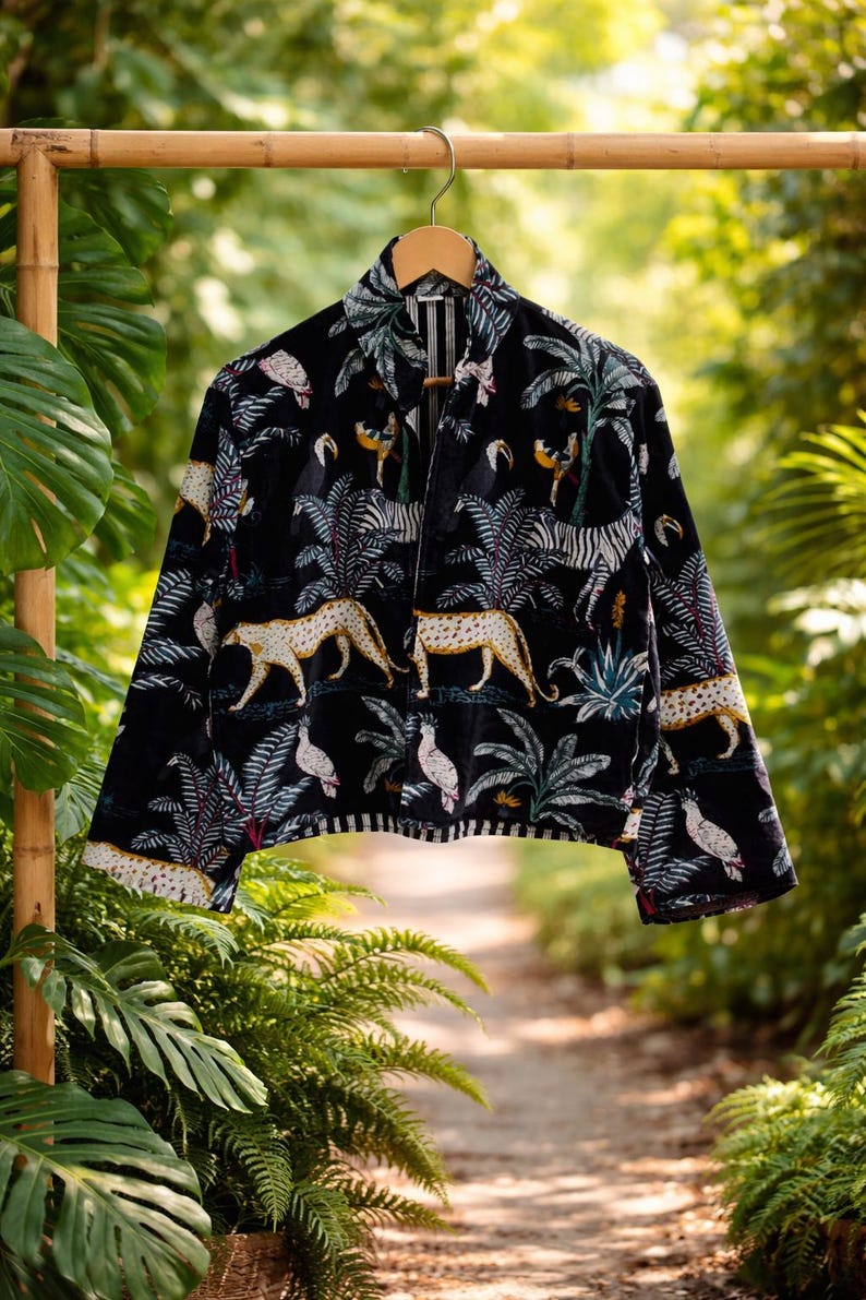 K&ouml;nnte beinhalten: Schwarze Samtjacke mit Dschungel-Print. Das Muster zeigt Leoparden, Zebras, V&ouml;gel und Palmen. Die Jacke h&auml;ngt an einem Holzb&uuml;gel vor dem Hintergrund &uuml;ppigen Gr&uuml;ns.