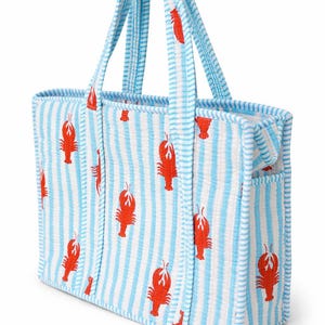 May include: A blue and white striped tote bag with a repeating pattern of red lobsters. The bag has two handles and a small pocket on the side. The bag is made of a textured fabric.
