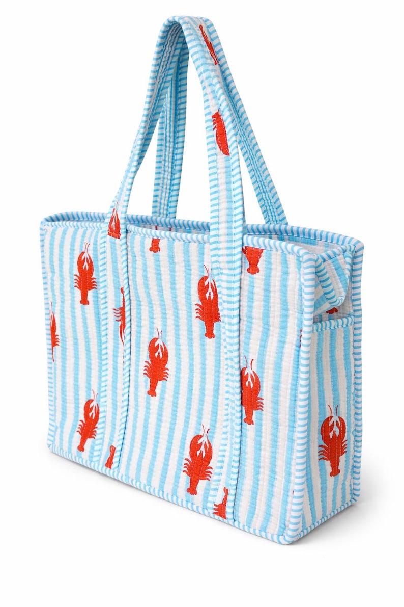 May include: A blue and white striped tote bag with a repeating pattern of red lobsters. The bag has two handles and a small pocket on the side. The bag is made of a textured fabric.