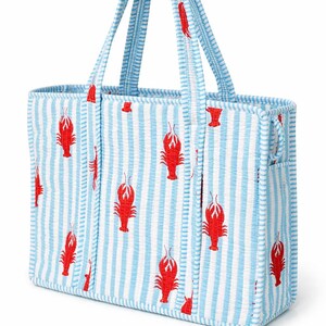 May include: A rectangular tote bag with blue and white vertical stripes and red lobster illustrations. The bag has matching striped handles and trim. The bag is made of a textured fabric.