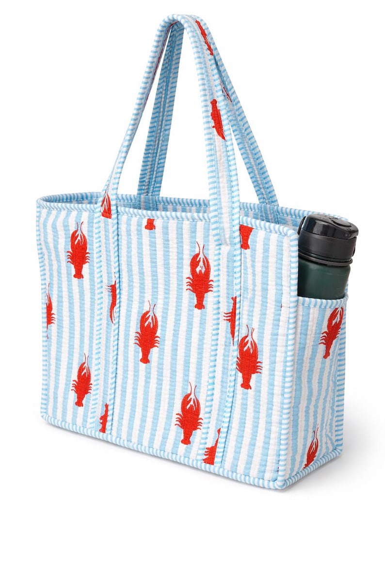 May include: A blue and white striped tote bag with a red lobster pattern. The bag has a pocket on the side and a green water bottle inside. The handles and trim are blue and white striped.
