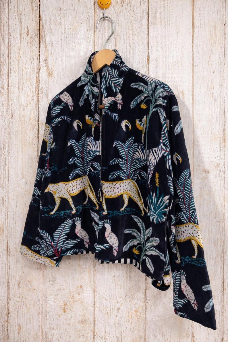 K&ouml;nnte beinhalten: Marineblaue Jacke mit Rei&szlig;verschluss und einem lebendigen Dschungel-Print mit Leoparden, Zebras, Papageien und Palmen. Die Jacke h&auml;ngt an einem Holzb&uuml;gel vor einer hellen Holzpaneelwand. Die Jacke hat einen hohen Kragen.