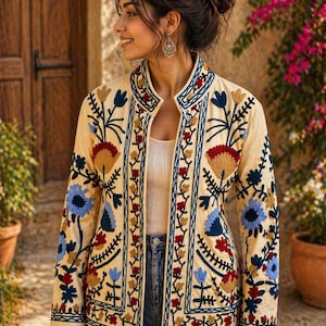 May include: Cream-colored embroidered jacket with a floral design in red, blue, and yellow. The jacket features a mandarin collar and is worn over a white top and blue jeans. The embroidery is detailed and covers the front and sleeves.