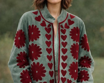 May include: A sage green jacket with a red floral and heart design. The jacket has a round collar and a red trim. The model is wearing a light green shirt and tan pants. The jacket is a unique piece of clothing.