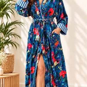 Op de afbeelding: Een lange, blauwe fluwelen kamerjas met een levendig bloemenpatroon met rode en gele bloemen. Het kledingstuk is voorzien van een bijpassende ceintuur in de taille, twee voorzakken en lange mouwen met blauw-witte gestreepte boorden.