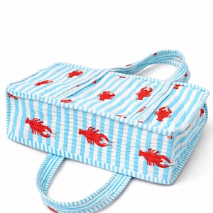 May include: A rectangular tote bag with a blue and white striped pattern and red lobster illustrations. The bag has two handles and is made of a textured fabric, perfect for carrying groceries or beach essentials.