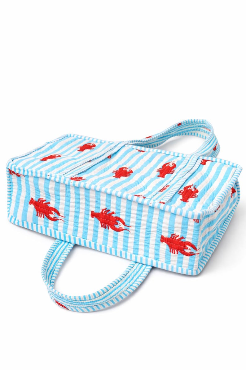 May include: A rectangular tote bag with a blue and white striped pattern and red lobster illustrations. The bag has two handles and is made of a textured fabric, perfect for carrying groceries or beach essentials.