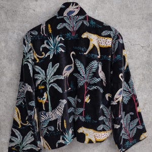 K&ouml;nnte beinhalten: Schwarze Samtjacke mit einem lebendigen Dschungel-Print mit Zebras, Leoparden, V&ouml;geln und &uuml;ppigem Laub. Die Jacke h&auml;ngt an einem silbernen Kleiderb&uuml;gel mit einem gelben Smiley.
