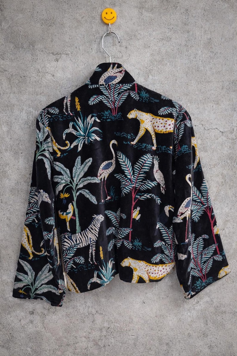 K&ouml;nnte beinhalten: Schwarze Samtjacke mit einem lebendigen Dschungel-Print mit Zebras, Leoparden, V&ouml;geln und &uuml;ppigem Laub. Die Jacke h&auml;ngt an einem silbernen Kleiderb&uuml;gel mit einem gelben Smiley.