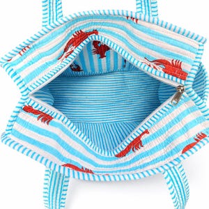 May include: Open view of a blue and white striped tote bag with red lobster print. The bag has a zipper closure and matching striped handles. The interior is also striped.