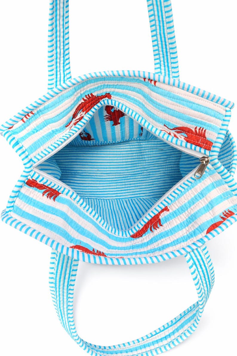 May include: Open view of a blue and white striped tote bag with red lobster print. The bag has a zipper closure and matching striped handles. The interior is also striped.