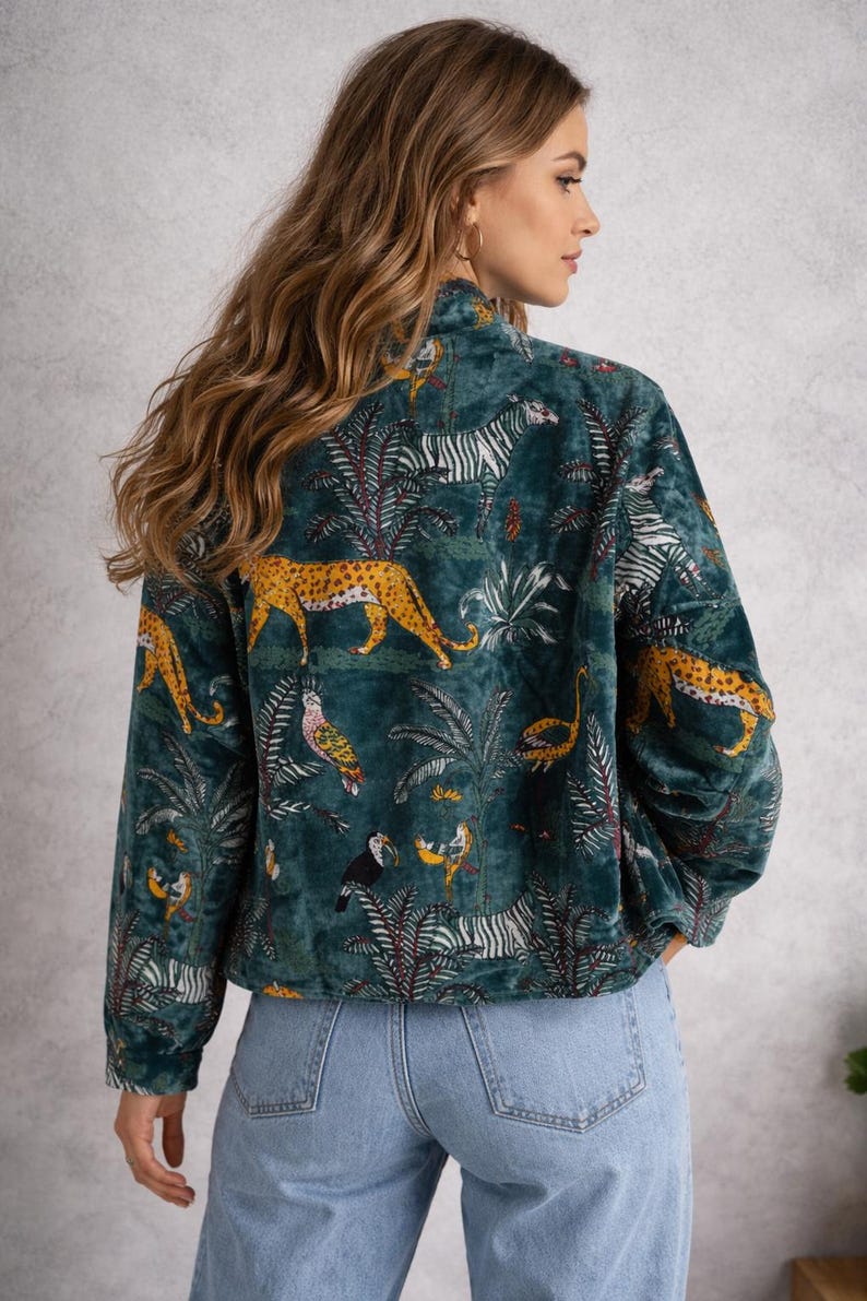 May include: Teal velvet jacket with a jungle-themed print. The design features zebras, leopards, birds, and tropical foliage. The jacket has a high collar and long sleeves, and is paired with blue jeans.